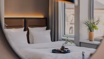 Et moderne, hyggeligt hotelværelse med pænt redte enkeltsenge, hvidt sengetøj, et stort vindue med beige gardiner og en bakke med morgenmadsprodukter på sengen. En vase med blomster står i vindueskarmen.