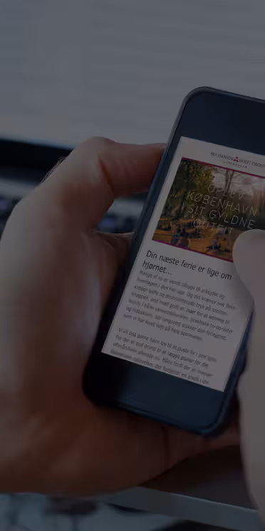 En person holder en smartphone, der viser en artikel, med en bærbar computer i baggrunden. Scenen foreslår multitasking og læsning på en digital enhed i kontormiljøer. Indholdet på telefonen er delvist synligt, men ulæseligt.