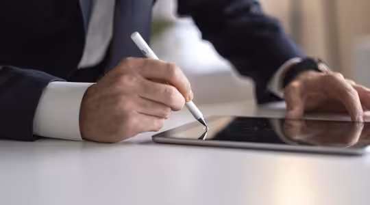 En person i et jakkesæt bruger en stylus til at skrive på en digital tablet, som hviler på et hvidt bord. Fokus er på hænderne med slørede baggrundselementer.