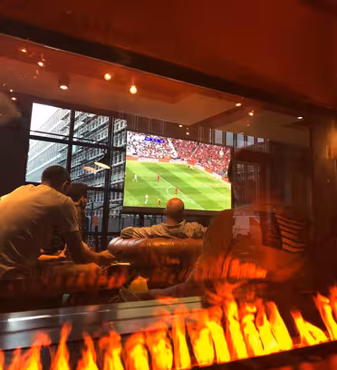 Folk i et hyggeligt lokale ser en fodboldkamp på et stort tv monteret på væggen. Scenen afspejles i et glaspanel, der viser levende flammer fra en pejs eller lignende installation, hvilket tilføjer varme til de moderne omgivelser.