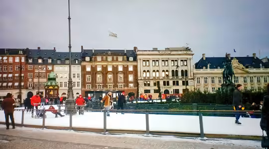 Folk, der skøjter på en udendørs skøjtebane på et bytorv, omgivet af historiske bygninger i europæisk stil. En statue og lygtepæl er synlige, og nogle mennesker ser på fra sidelinjen. Det ser ud til at være dagtimerne.
