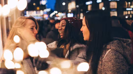 Tre kvinder smiler og taler i en festlig, oplyst gade om natten. De er iført vintertøj, inklusive frakker og en rød hue. Sløret lys skaber en varm forgrund, der forstærker den muntre, hyggelige atmosfære.