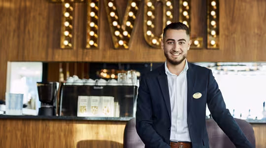 En mand i jakkesæt står smilende foran et godt oplyst skilt, hvor der står "Tivoli". Bag ham står en disk med kaffemaskine og menuer. Rammerne ser ud til at være en stilfuld café eller restaurant.