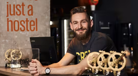 En smilende mand med skæg læner sig op ad en disk i receptionen på et hostel. Adskillige gear-formede priser vises foran ham. Baggrunden har teksten "vi er bare et herberg" i orange.
