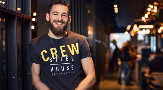 Mand med smilende skæg, iført en "CREW STEEL HOUSE COPENHAGEN"-t-shirt, står afslappet op ad en mørk væg i et varmt oplyst interiør, muligvis en café eller restaurant, med slørede mennesker i baggrunden.