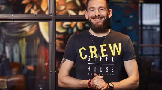 En skægget mand med et venligt smil står indendørs, iført en sort T-shirt med gul tekst, hvor der står "CREW STEEL HOUSE COPENHAGEN." Baggrunden byder på farverig, abstrakt vægkunst.