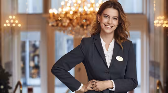 En smilende person i et professionelt jakkesæt står indendørs med en lysekrone og store vinduer i baggrunden, hvilket tyder på et formelt mødested som et hotel eller et konferencecenter.