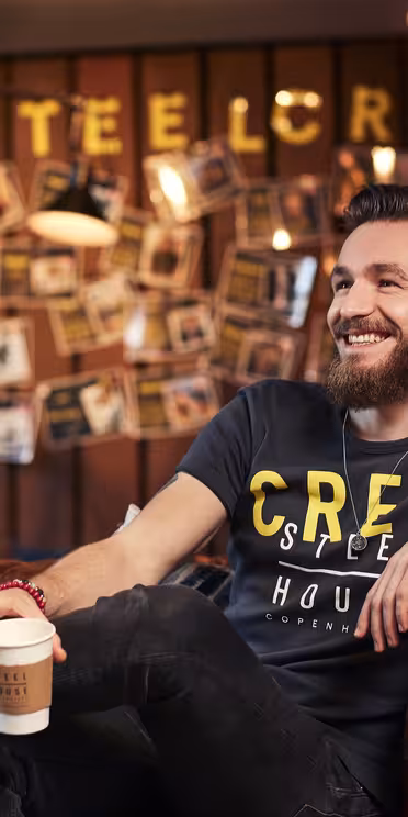En mand med skæg sidder på en brun lædersofa, holder en kaffekop og smiler. Han har en mørk t-shirt på med "CREW" trykt på. Baggrunden har en væg med fotos og "#STEELCREW" med gule bogstaver.