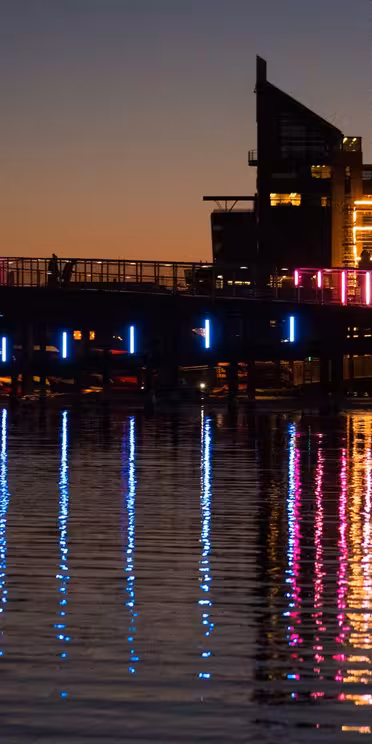 En bro over vand i skumringen, oplyst med farverige neonlys, der reflekterer på vandoverfladen. Silhouettede bygninger og en byggekran er synlige mod den mørkere himmel.