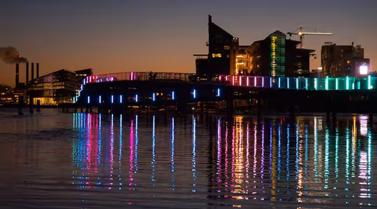 En bro over vand i skumringen, oplyst med farverige neonlys, der reflekterer på vandoverfladen. Silhouettede bygninger og en byggekran er synlige mod den mørkere himmel.