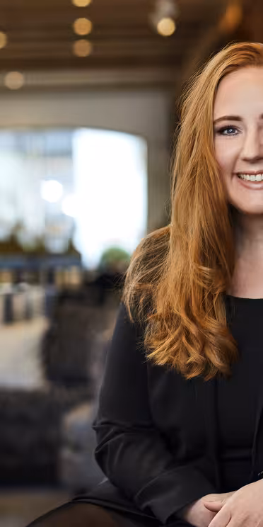 En person med langt rødt hår, iført en sort blazer, sidder og smiler i stilfulde indendørs omgivelser med moderne indretning. Stearinlys og puder er synlige i den blødt oplyste baggrund.
