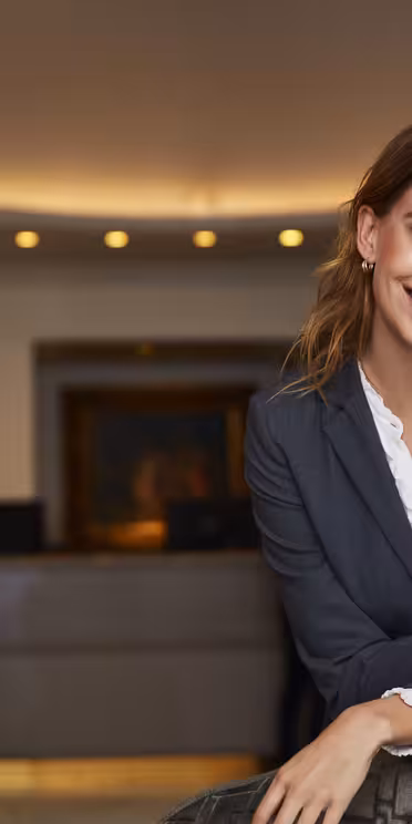 En smilende person i et formelt jakkesæt læner sig op ad et bord i en hotellets lobby. Baggrunden har en reception med en medarbejder og blød overheadbelysning, hvilket skaber en indbydende atmosfære.