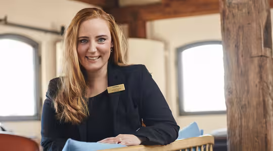 En kvinde med langt rødt hår iført en sort blazer og navneskilt smiler, mens hun sidder indendørs. Hun læner sig op af en blå pude og et trærækværk med solbeskinnede buede vinduer og træbjælker i baggrunden.