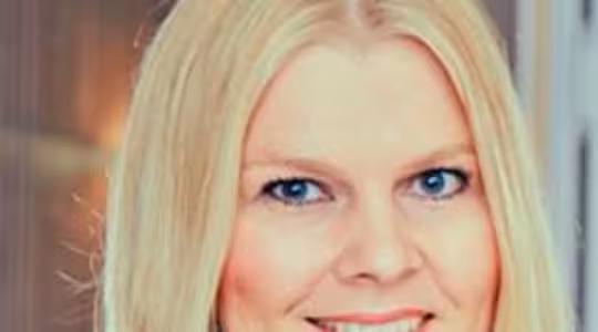 En person med langt blondt hår, der smiler til kameraet, iført en blå top. Baggrunden ser ud til at være indendørs med slørede elementer og blød belysning.