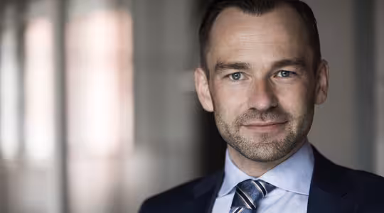 En mand i jakkesæt og slips står i et blødt oplyst indendørs miljø. Han har kort hår og trimmet skæg med et lille smil. Baggrunden er sløret, hvilket tyder på et professionelt miljø.