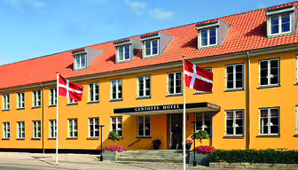 Billedet viser en gul bygning med rødt tegltag, identificeret som Gentofte Hotel. To danske flag er vist foran. Indgangen er flankeret af potteplanter og trapper, der fører op til døren, sat mod en klar blå himmel.