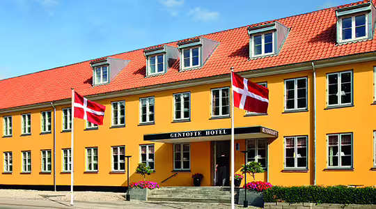 Billedet viser en gul bygning med rødt tegltag, identificeret som Gentofte Hotel. To danske flag er vist foran. Indgangen er flankeret af potteplanter og trapper, der fører op til døren, sat mod en klar blå himmel.