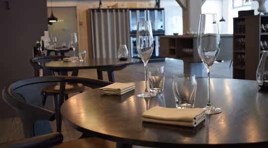 Restaurantinteriør med moderne træborde sat med hvide servietter, vandglas og champagnefløjter. Blød belysning og træbjælker skaber en hyggelig atmosfære. Tomme stole foreslår en klar, men ubesat spiseplads.
