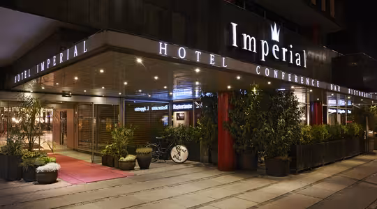 Indgang til Imperial Hotel med lysskilte til hotel, konference, bar og restaurant. Det ydre er beklædt med planter og har en rød løber, der fører til indgangen. Et skilt til cykling er synligt i nærheden.