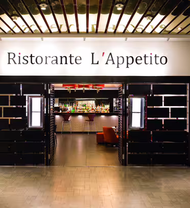 Indgang til "Ristorante L'Appetito" med et moderne interiør med en bar synlig gennem åbne dobbeltdøre. Designet omfatter mørke træelementer og diskret belysning, hvilket skaber en indbydende atmosfære.