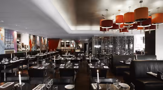 Et moderne restaurantinteriør med mørke træborde, sorte siddepladser og røde accenter. Værelset har en væg af vinflasker, farverige kunstværker, elegante lysekroner og borde dækket med glasvarer og stearinlys.