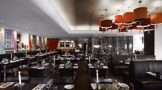Moderne restaurantinteriør med mørke træborde sat med glasvarer og hvide servietter. Lædersæder, røde lysekroner og farverig vægkunst forbedrer stemningen. En vinreol er synlig i baggrunden.