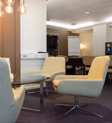 Et moderne loungeområde med cremefarvede stole omkring et glasbord, svagt oplyst af hængende pærer. I baggrunden er der flere stole og et flipover, sat op mod en neutral tonet væg og loft.