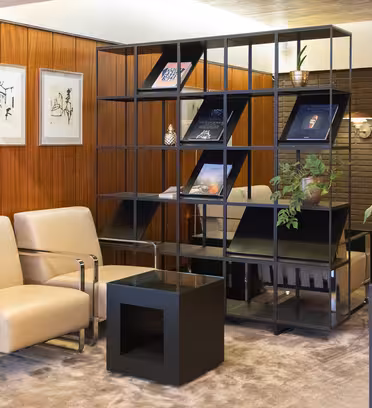 Moderne interiør med to beige lænestole, en sort metalhylde med kunst og bøger og en stor keramikkrukke med grønt. Rummet har træpaneler, indrammet kunst på væggen og omgivende belysning, der skaber en hyggelig atmosfære.