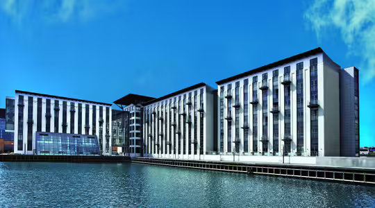 Moderne bygning ved vandet med høje, lodrette vinduer og altaner ud mod et vandområde. Strukturen har et symmetrisk design og er sat mod en klar blå himmel.