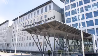 Moderne bygning af Hotel Copenhagen Island med minimalistisk design. Funktionerne omfatter en glasindgang understøttet af metalbjælker og store vinduer. Hotellets navn er tydeligt vist nær indgangen.