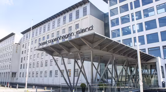 Moderne bygning af Hotel Copenhagen Island med minimalistisk design. Funktionerne omfatter en glasindgang understøttet af metalbjælker og store vinduer. Hotellets navn er tydeligt vist nær indgangen.