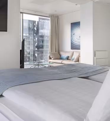 Moderne hotelværelse med en pænt lavet dobbeltseng med hvidt sengetøj og et gråt sengetøj. Et fladskærms-tv er monteret på væggen. Store vinduer afslører en byudsigt med moderne bygninger. En hyggelig sofa og abstrakt kunst dekorerer rummet.