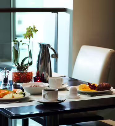 Et moderne morgenbord med hvide stole. Den byder på tallerkener med skåret frugt, korn, røræg og en rundstykke sammen med en kop kaffe, et kogt æg og en karaffel. En potteplante og krydderier pryder bordet.