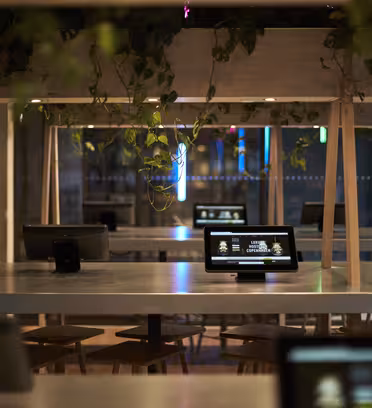 Et moderne cafeinteriør med lange fællesborde, hver udstyret med digitale tablets. Hængende planter pryder loftet. Atmosfæren er blødt oplyst, hvilket skaber en hyggelig og teknologisk stemning, med et neonskilt synligt i baggrunden.