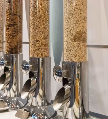 Række af korndispensere på en bordplade, hver fyldt med forskellige typer korn, inklusive flager og granola. Dispenserne har metalbunde og klare beholdere, så indholdet kan ses.