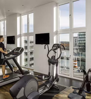 En person bruger et løbebånd i et lyst moderne fitnesscenter med store vinduer med udsigt over bybilledet og vandet. I nærheden er en ellipsemaskine og en romaskine. To vægmonterede tv er synlige. Planter dekorerer rummet.