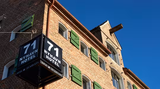 Murstensbygning under klar blå himmel med et hjørneskilt, hvor der står "71 NYHAVN HOTEL." Bygningen har grønne vinduesskodder og en markant, traditionel arkitektonisk stil.