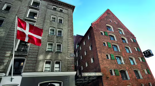 Gadebillede af en historisk bygning, der huser "71 Nyhavn Hotel", med murstens- og stenfacader. Et dansk flag er i forgrunden, og himlen er klarblå, hvilket tilføjer den maleriske scene.