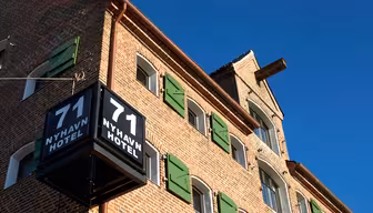 Udsigt over en murstensbygning med flere vinduer med grønne skodder. På et fremtrædende skilt står "71 Nyhavn Hotel" med hvid tekst på sort baggrund. Himlen er klar og blå.
