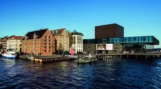 Panoramaudsigt over havnefronten i København. Historiske murstensbygninger ligger langs venstre side, mens en moderne glas- og murstenskonstruktion rager ud over vandet. Bådene ligger til kaj langs marinaen under en klar blå himmel.