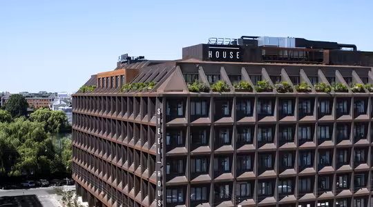 En stor, moderne bygning mærket "Stålhus" med en geometrisk facade og grønne områder på taget. Det er sat mod en klar blå himmel, med omkringliggende træer og bybilledet i baggrunden.