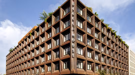 En moderne bygning i flere etager med et geometrisk design og rustfarvet facade under en blå himmel. Bygningen har store vinduer og grønne områder på taget. Cykler og et køretøj er parkeret i nærheden, med skiltning med "Stålhus" på hjørnet.
