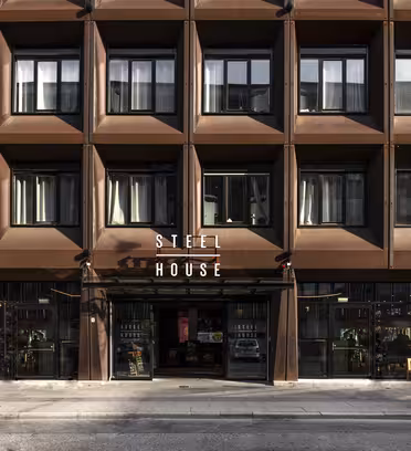 En moderne bygningsfacade med et gitterlignende design af store vinduer og brune rammer. Indgangen har ordene "Stålhus" over glasdøre. Bænke og potteplanter er placeret langs fortovet.