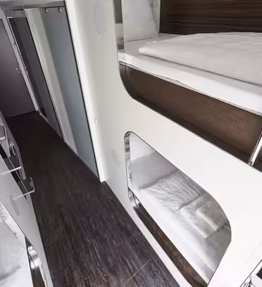 Moderne, smal gang med køjesenge i pod-stil med slanke, hvide paneler på begge sider. Det træteksturerede gulv tilføjer varme, mens forsænket belysning forbedrer det moderne design. En lukket dør er synlig for enden af gangen.