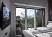 Et moderne soveværelse med en pæn opredt seng, store glasskydedøre og en udendørs udsigt til grønt og et rækværk. Et fladskærms-tv er monteret på væggen til venstre, og sollys oplyser rummet.