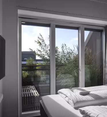 Et moderne soveværelse med en pæn opredt seng, store glasskydedøre og en udendørs udsigt til grønt og et rækværk. Et fladskærms-tv er monteret på væggen til venstre, og sollys oplyser rummet.