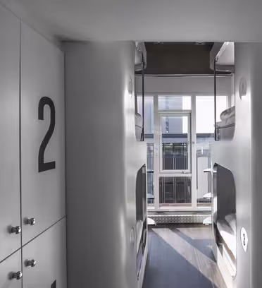 En moderne kollegiegang med nummererede skabe til venstre og indbyggede køjesenge til højre. Sollyset filtrerer gennem et vindue for enden og oplyser det slanke, minimalistiske design.