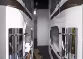 En gang på et moderne kapselhotel med slanke, hvide soveposer stablet på begge sider. Hver pod har en skinnende metallisk facade, og rummet har et mørkt trægulv. Gangen fører til en lys døråbning for enden.