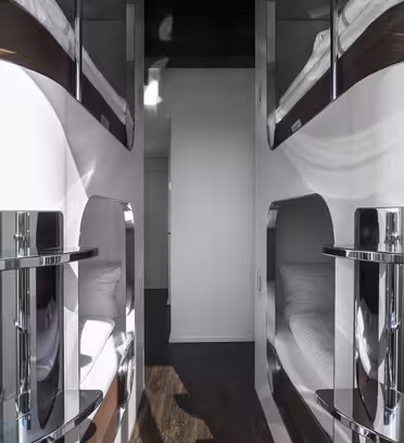 En gang på et moderne kapselhotel med slanke, hvide soveposer stablet på begge sider. Hver pod har en skinnende metallisk facade, og rummet har et mørkt trægulv. Gangen fører til en lys døråbning for enden.