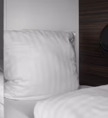 En lille hyggelig sovepose med en blød hvid pude og sengetøj. Væggene er glatte, og der er et mørkt træpanel med påsat en sort lomme. Et nummer 1 er synligt på partitionen. Atmosfæren er minimalistisk og behagelig.